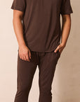 A man wearing a matching dark brown short-sleeve t-shirt and jogger pants, standing against a neutral beige background.