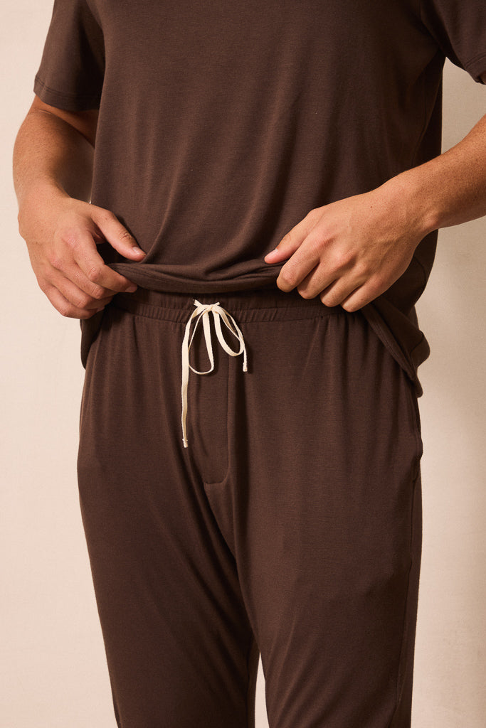 A close-up of a man wearing a matching brown lounge set, featuring a short-sleeve top and drawstring pants, with their hands pulling the waistband of the pants slightly outward.