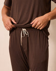 A close-up of a man wearing a matching brown lounge set, featuring a short-sleeve top and drawstring pants, with their hands pulling the waistband of the pants slightly outward.