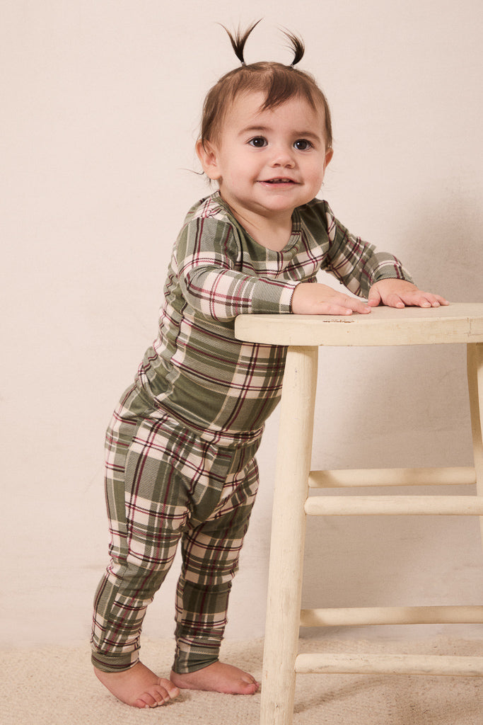 A toddler standing barefoot and leaning on a light wooden stool, wearing a long-sleeve plaid pajama set in muted green, beige, and burgundy tones against a plain cream backdrop.