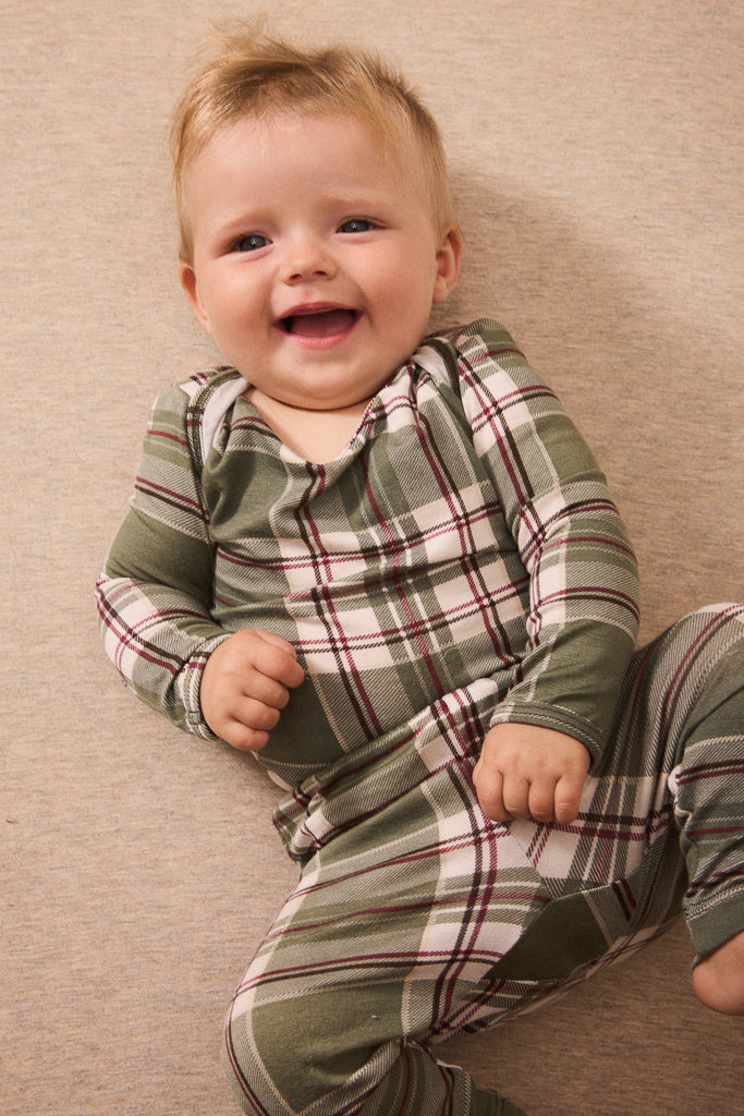 A smiling baby lying on a beige carpet, wearing a green and white plaid long-sleeve pajama top and matching pants.