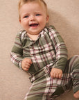 A smiling baby lying on a beige carpet, wearing a green and white plaid long-sleeve pajama top and matching pants.