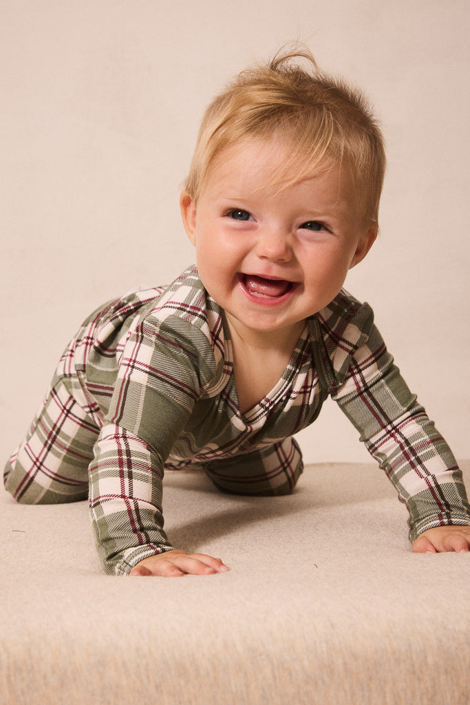 A smiling baby crawling on a beige fabric surface, wearing a long-sleeve plaid pajama top in muted green, beige, and burgundy.