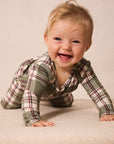 A smiling baby crawling on a beige fabric surface, wearing a long-sleeve plaid pajama top in muted green, beige, and burgundy.