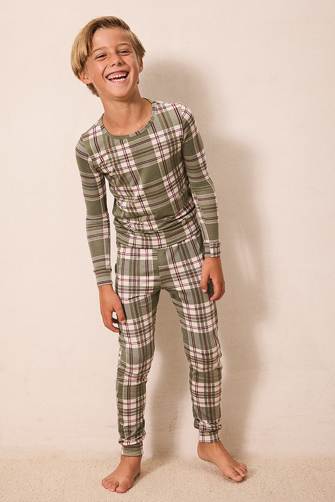 A young boy wearing a matching long-sleeve plaid pajama set in olive green, pink, and white, standing against a plain beige wall.
