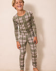 A young boy wearing a matching long-sleeve plaid pajama set in olive green, pink, and white, standing against a plain beige wall.