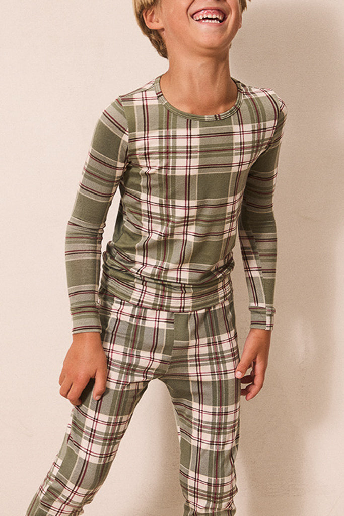 A child wearing a matching long-sleeve top and pants pajama set in an olive green plaid pattern with beige and white stripes, set against a plain beige wall.