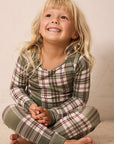 A young blonde girl sitting cross-legged on a beige carpet, wearing a green, white, and burgundy plaid long-sleeve pajama set.