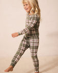A young blonde girl wearing a green and pink plaid long-sleeve pajama set, captured mid-step against a warm beige studio backdrop.