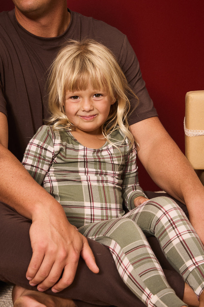 A young blonde girl wearing a green and red plaid pajama set sits on her father's lap against a dark red background.