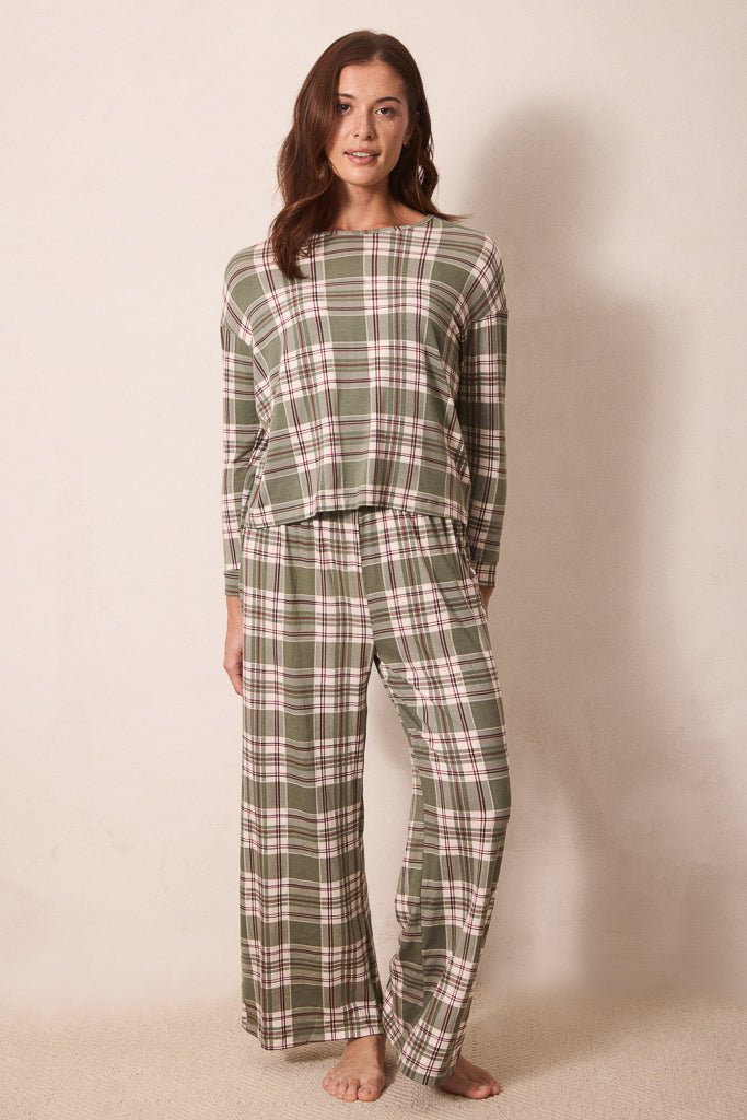 A woman modeling a two-piece plaid pajama set consisting of a long-sleeve top and matching wide-leg pants in an olive green, pink, and white plaid pattern, standing against a plain beige wall.