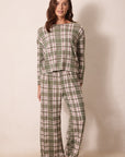 A woman modeling a two-piece plaid pajama set consisting of a long-sleeve top and matching wide-leg pants in an olive green, pink, and white plaid pattern, standing against a plain beige wall.