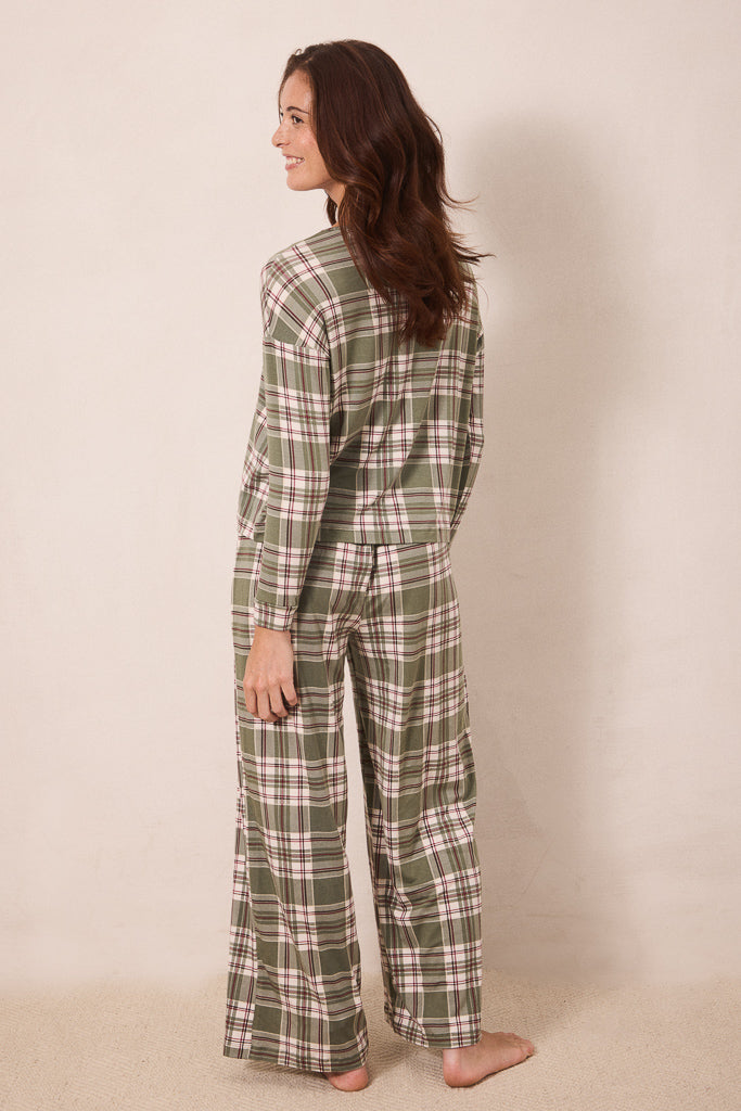 A woman wearing a matching long-sleeve plaid pajama set in sage green, cream, and white, shown from a side-back angle against a plain beige studio backdrop.