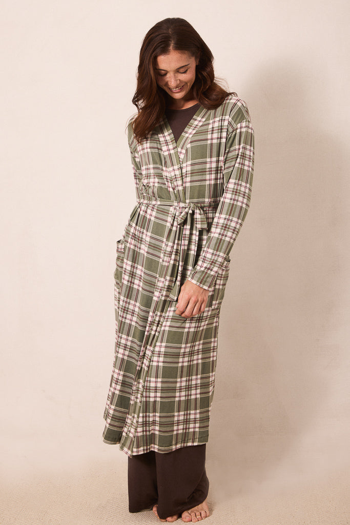 A woman modeling a long-sleeve plaid bathrobe in sage green, cream, and brown, standing against a plain beige studio background.