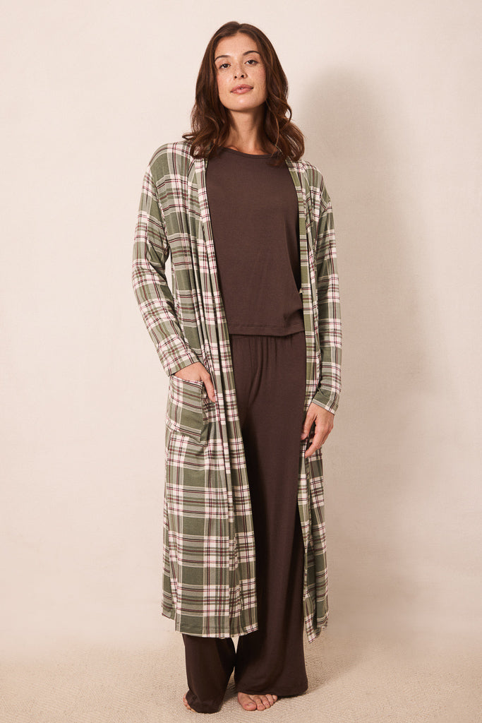 A woman stands barefoot against a plain beige studio background wearing a long green plaid robe over a dark brown lounge set.