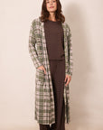 A woman stands barefoot against a plain beige studio background wearing a long green plaid robe over a dark brown lounge set.