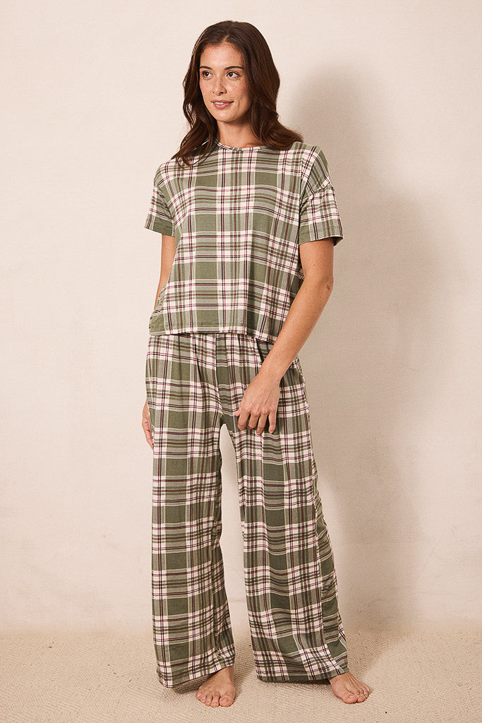 A woman modeling a two-piece plaid pajama set consisting of a short-sleeve top and matching wide-leg pants in an olive green, beige, and white plaid pattern, standing against a plain beige wall.