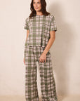 A woman modeling a two-piece plaid pajama set consisting of a short-sleeve top and matching wide-leg pants in an olive green, beige, and white plaid pattern, standing against a plain beige wall.