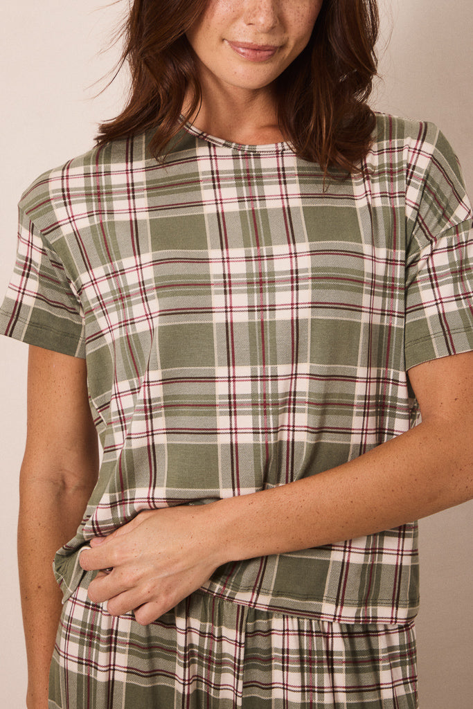 A woman wearing a short-sleeve plaid top in sage green, cream, and burgundy, shown from the shoulders to the waist against a plain beige background.