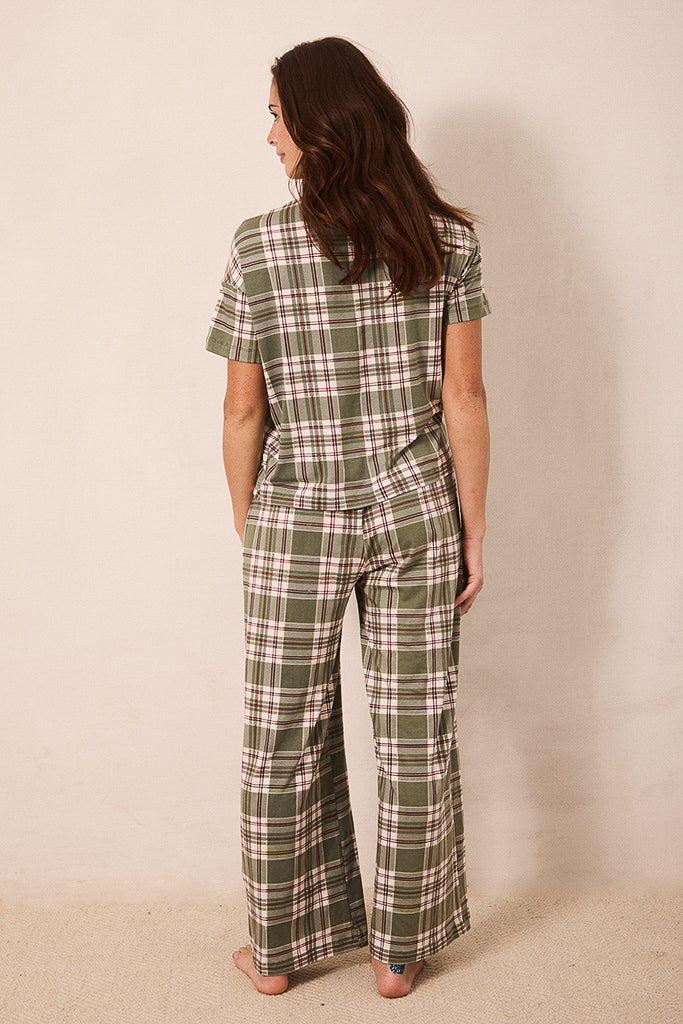 A woman shown from the back wearing a short-sleeve plaid jumpsuit in olive green, beige, and white, standing against a plain beige wall.