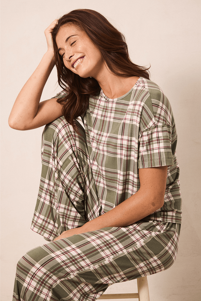 A woman wearing a matching plaid pajama set with a short-sleeve top and pants, seated on a stool against a plain beige background.