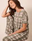 A woman wearing a matching plaid pajama set with a short-sleeve top and pants, seated on a stool against a plain beige background.