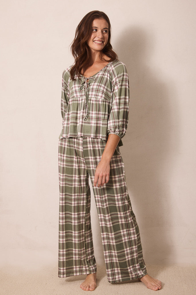 A woman modeling a two-piece plaid pajama set consisting of a long-sleeve top with a tie neckline and matching wide-leg pants, in an olive green, beige, and white plaid pattern against a plain beige studio background.