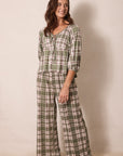 A woman modeling a two-piece plaid pajama set consisting of a long-sleeve top with a tie neckline and matching wide-leg pants, in an olive green, beige, and white plaid pattern against a plain beige studio background.