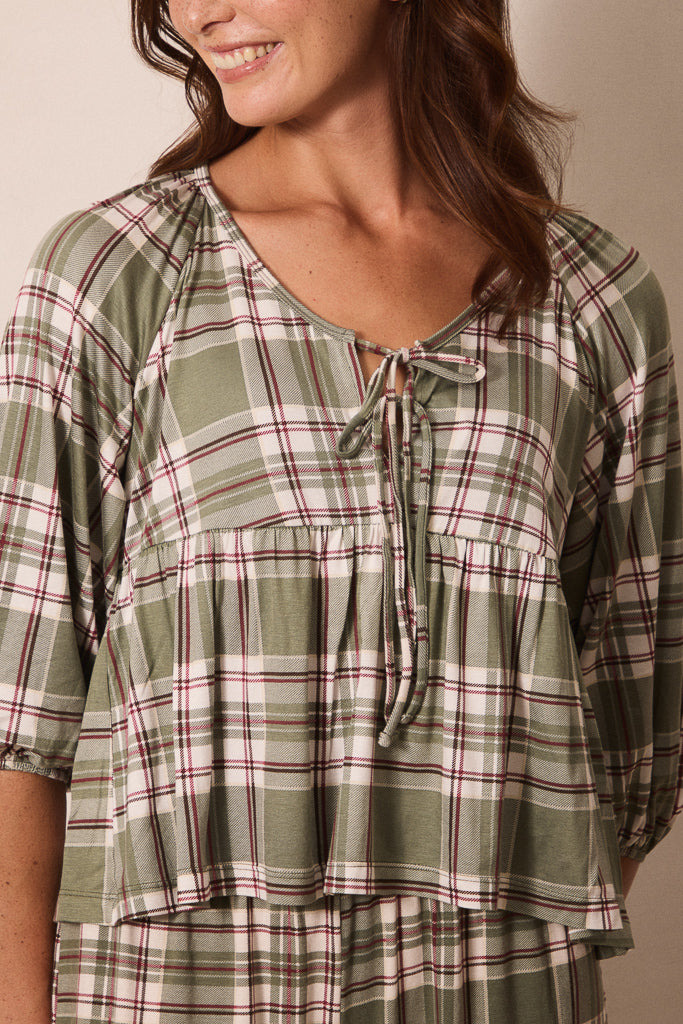 A woman wearing a green plaid short-sleeve top with a tie neckline, shown in a close-up shot against a neutral beige background.
