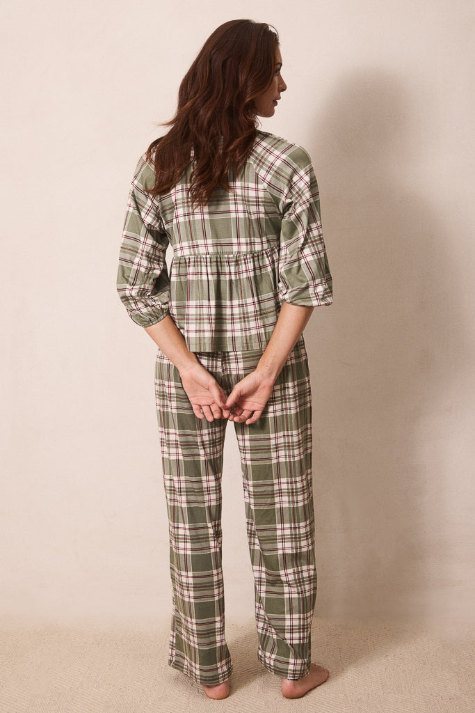 A woman stands barefoot against a plain beige wall, viewed from the back, wearing a green plaid pajama set with a long-sleeve top and matching pants.