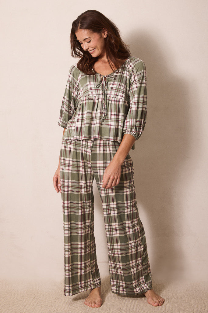 A woman modeling a two-piece plaid pajama set consisting of a long-sleeve top with a tie neckline and matching wide-leg pants, in an olive green, beige, and white plaid pattern against a plain beige background.
