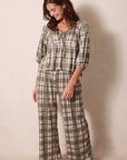 A woman modeling a two-piece plaid pajama set consisting of a long-sleeve top with a tie neckline and matching wide-leg pants, in an olive green, beige, and white plaid pattern against a plain beige background.