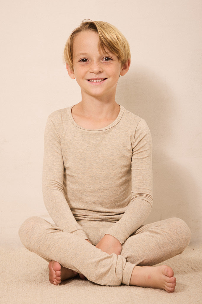 A young boy with blonde hair sits cross-legged on a light beige carpet, wearing a matching beige long-sleeve top and pants set against a plain beige wall.