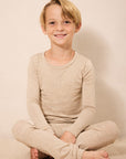 A young boy with blonde hair sits cross-legged on a light beige carpet, wearing a matching beige long-sleeve top and pants set against a plain beige wall.