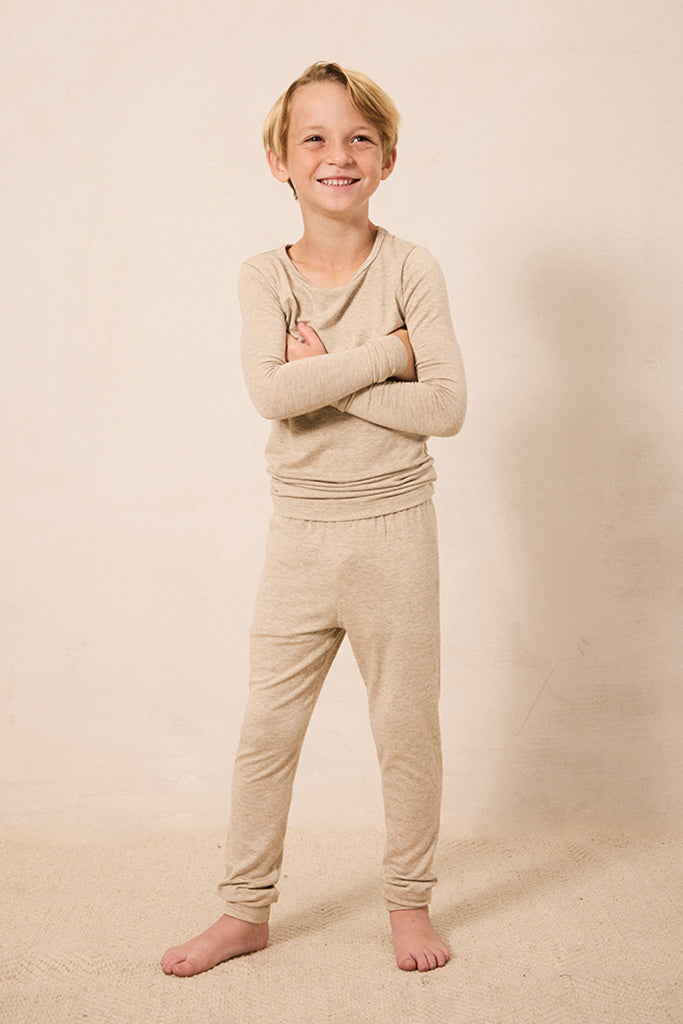A young boy with blonde hair stands barefoot against a warm beige studio backdrop, wearing a matching beige long-sleeve top and jogger-style pants set.
