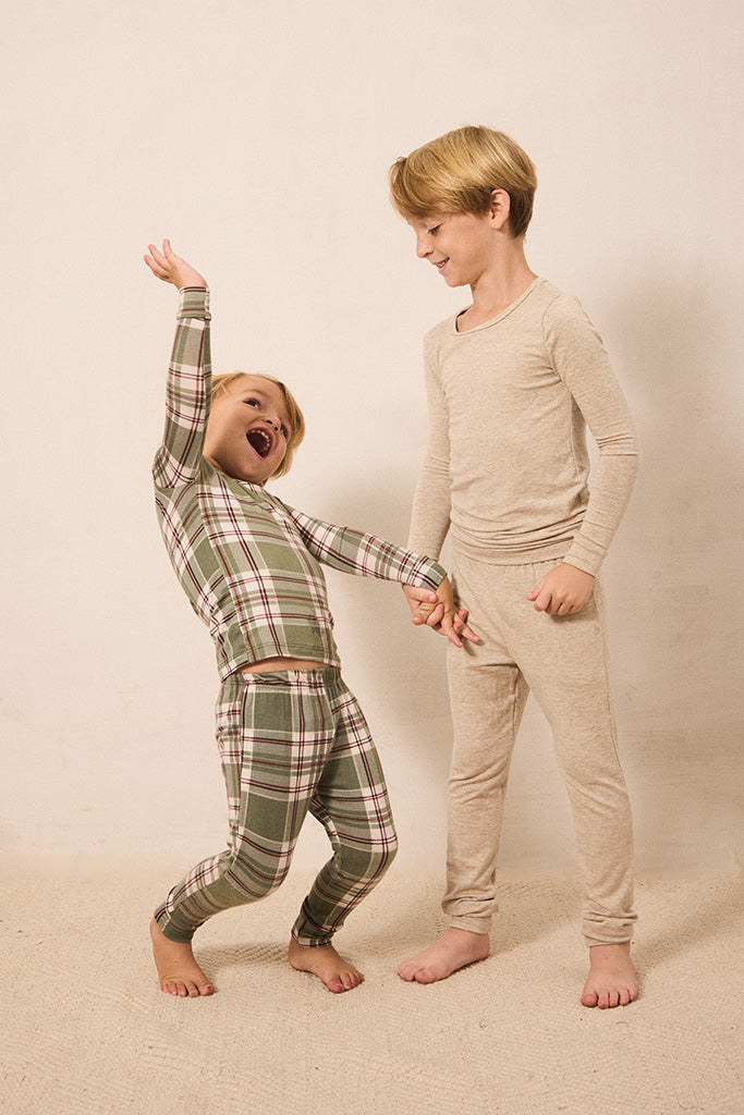 Two children modeling long-sleeve pajamas in a studio setting. The child on the left wears a green, pink, and white plaid pajama set and is dancing joyfully while holding hands with the child on the right, who wears a solid beige pajama set.