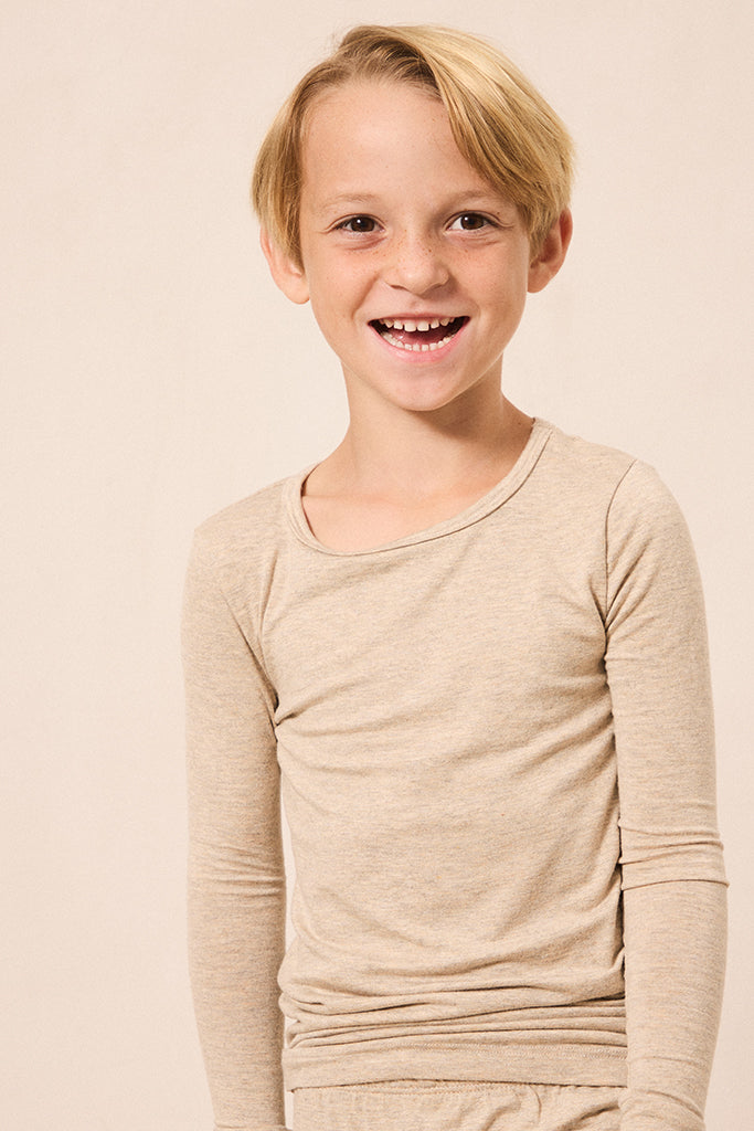 A young blonde boy smiling, wearing a light beige long-sleeve crewneck shirt against a solid light beige background.