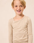A young blonde boy smiling, wearing a light beige long-sleeve crewneck shirt against a solid light beige background.