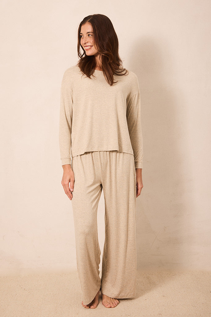 A woman modeling a matching beige knit loungewear set consisting of a long-sleeve cropped top and wide-leg pants, standing against a plain beige studio backdrop.