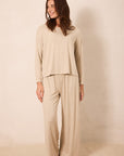 A woman modeling a matching beige knit loungewear set consisting of a long-sleeve cropped top and wide-leg pants, standing against a plain beige studio backdrop.