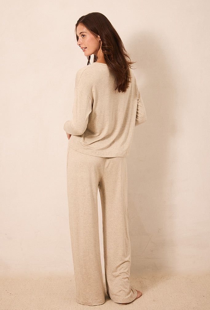 A woman stands barefoot against a plain beige wall, wearing a matching beige knit lounge set consisting of a long-sleeve top and wide-leg pants.