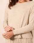 A woman wearing a light beige long-sleeve knit top and matching bottoms, shown from the shoulders to the hips against a plain warm beige background.