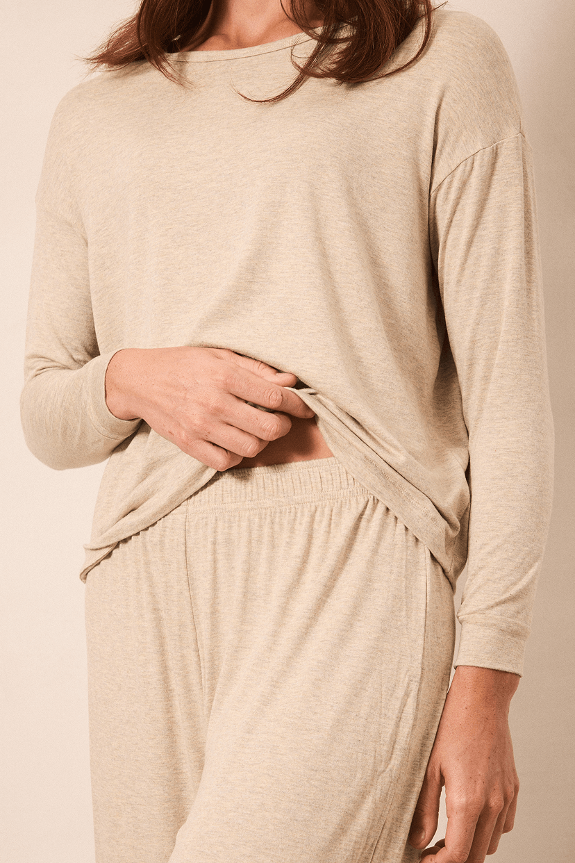A woman wearing a light beige long-sleeve knit top and matching lounge pants, shown from the shoulders to the upper thighs against a plain warm beige studio background.