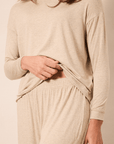 A woman wearing a light beige long-sleeve knit top and matching lounge pants, shown from the shoulders to the upper thighs against a plain warm beige studio background.