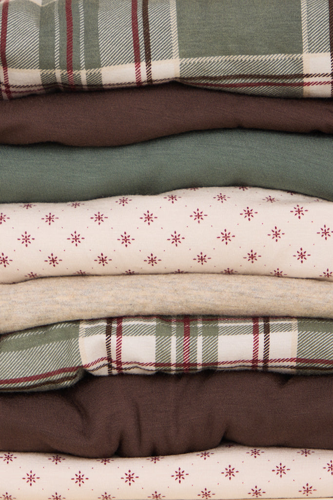 A stacked collection of folded fabrics in various patterns, including plaid and small star prints, in muted earthy tones of green, brown, beige, and cream.