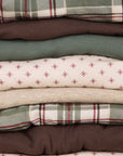 A stacked collection of folded fabrics in various patterns, including plaid and small star prints, in muted earthy tones of green, brown, beige, and cream.
