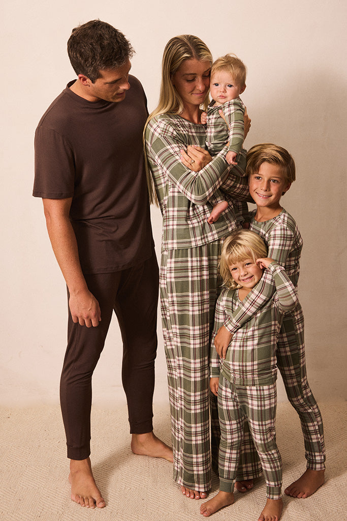 A family of five wearing matching plaid pajamas stands together against a plain beige studio background. The father wears a solid dark brown pajama set, while the mother, baby, and two children wear green, beige, and brown plaid long-sleeve pajama sets.