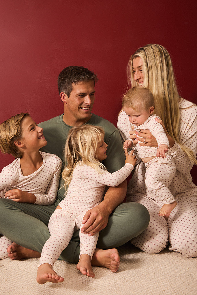 A family of five wearing matching light red and white twinkle star pajamas sits together on a cream carpet against a solid dark red background.