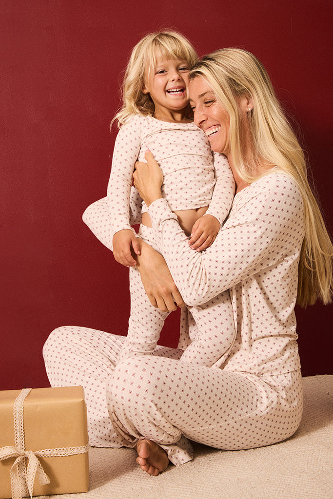 A smiling blonde woman and young blonde child wearing matching cream pajamas with small red polka dots, sitting together against a dark red background with a wrapped gift box on the floor.