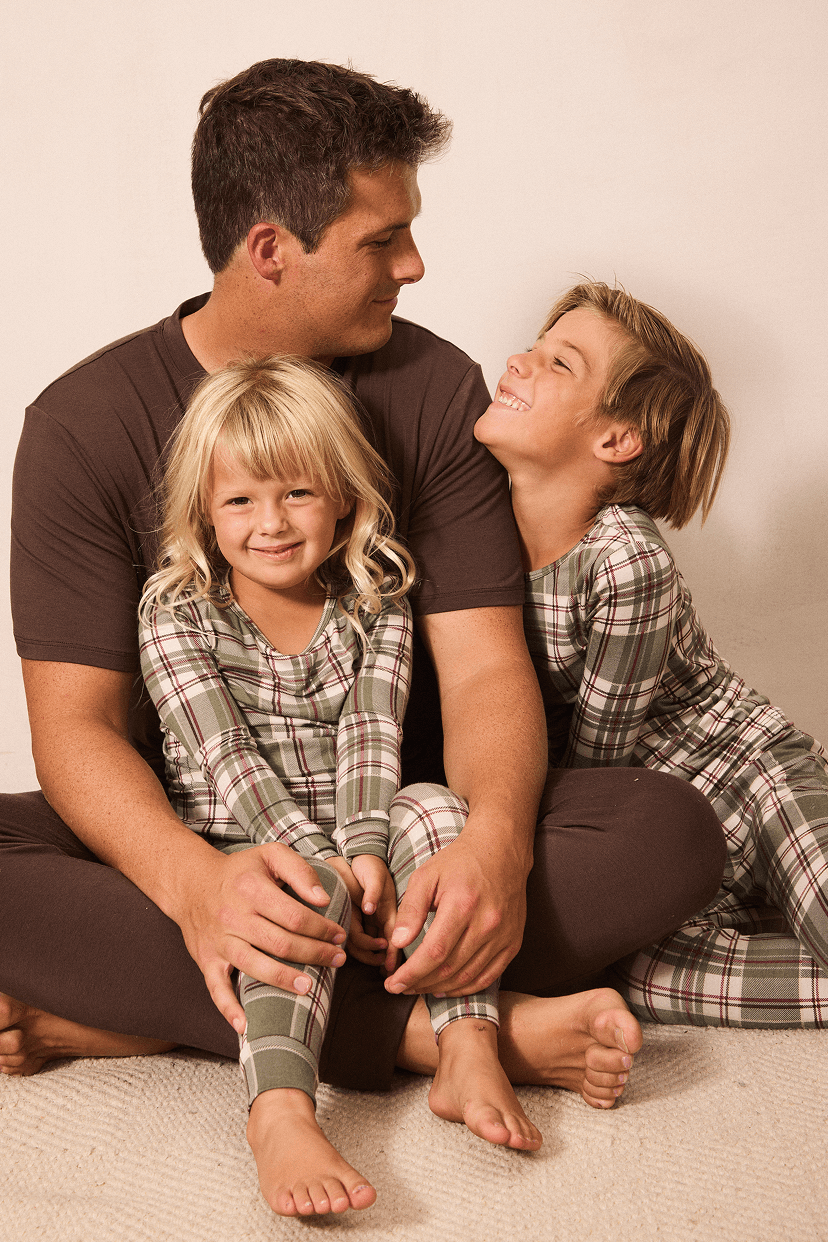A father and two children wearing matching brown and green plaid pajamas sitting together on a light beige floor against a plain beige wall.